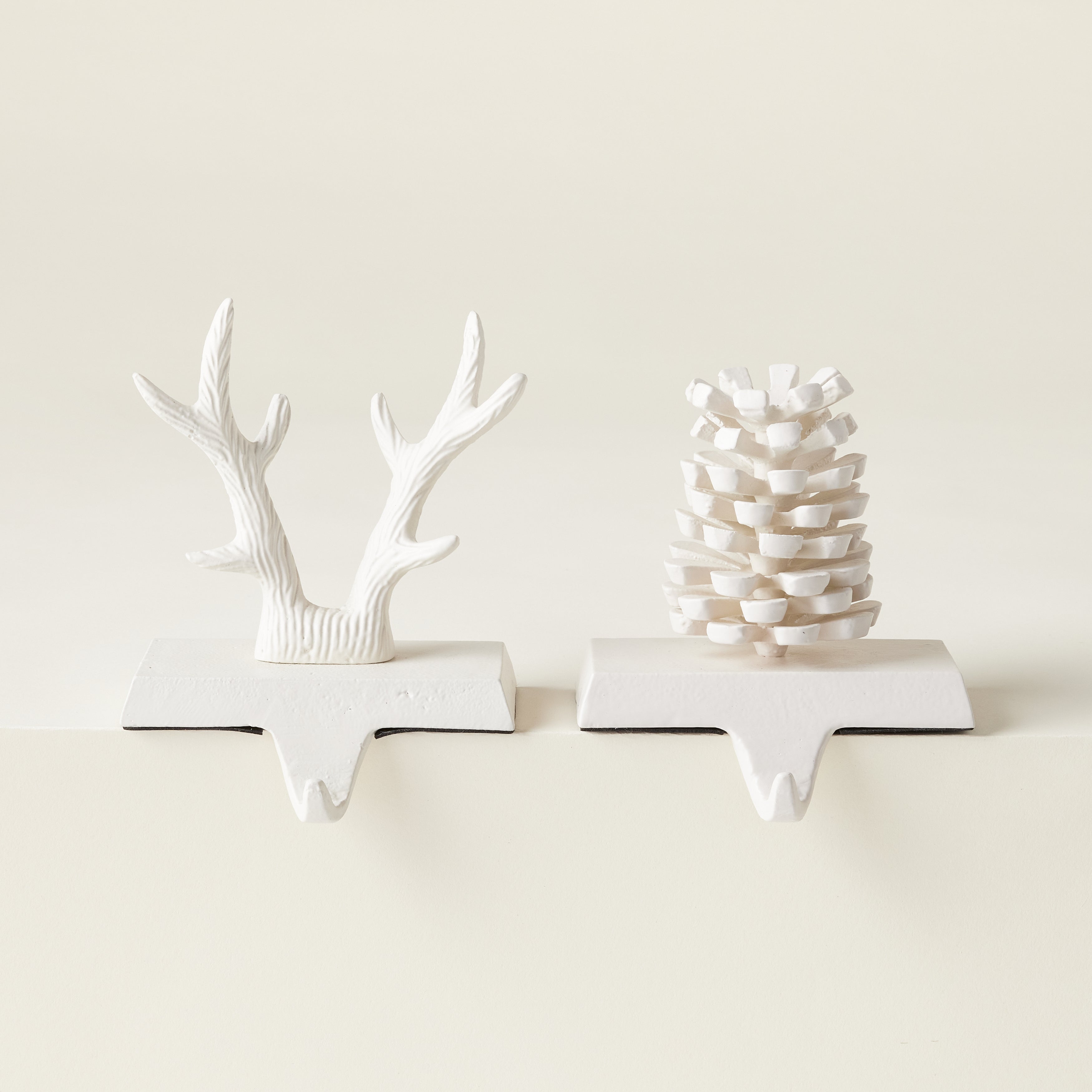 A set of two matte white iron stocking holders, one shaped like an acorn and the other like reindeer antlers.