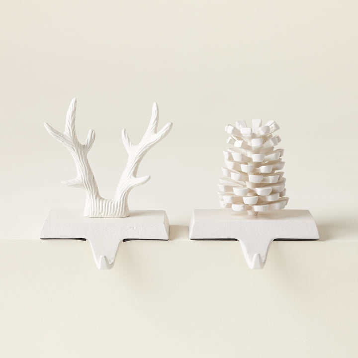A set of two matte white iron stocking holders, one shaped like an acorn and the other like reindeer antlers.