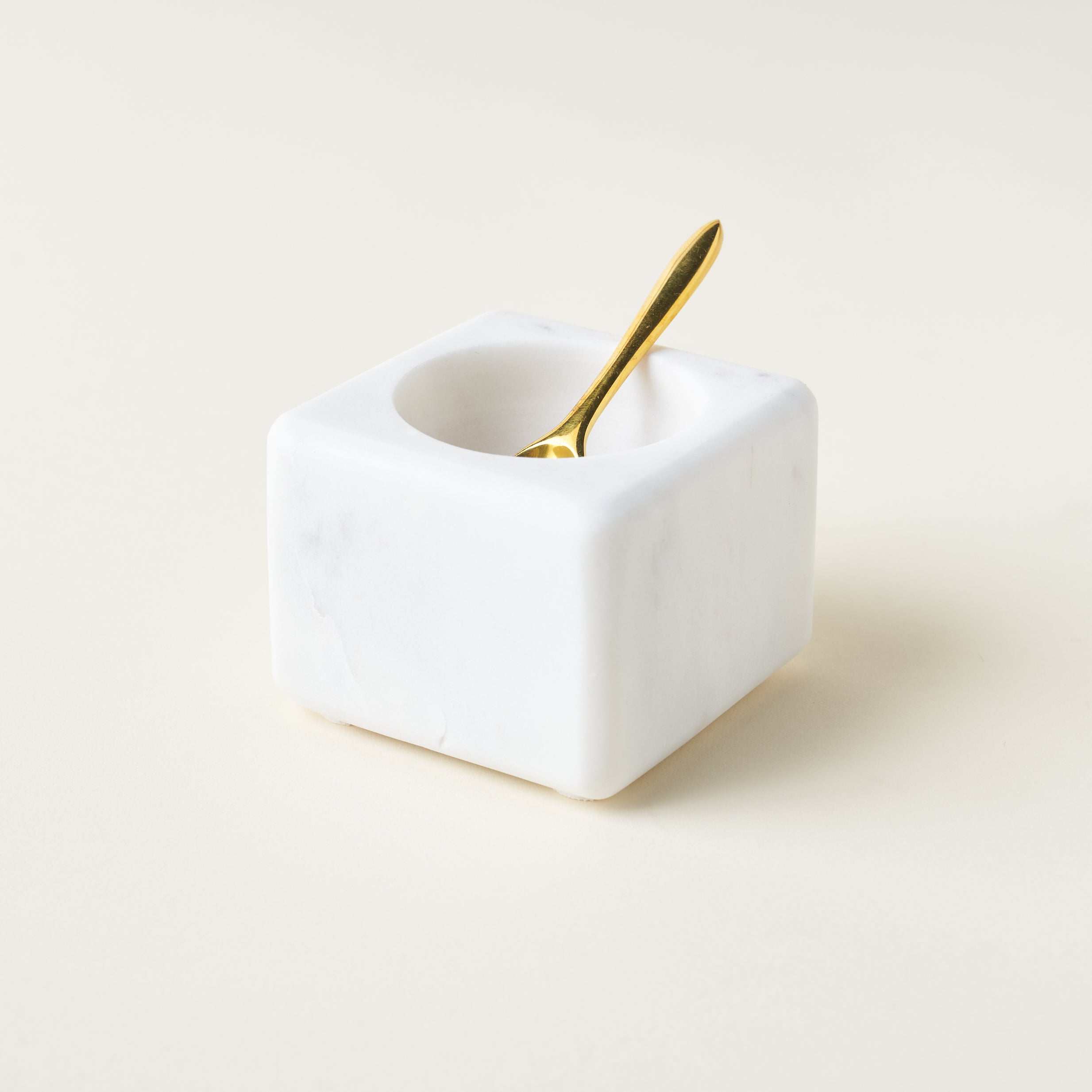 A square, white marble bowl with a rounded opening, accompanied by a polished brass spoon.
