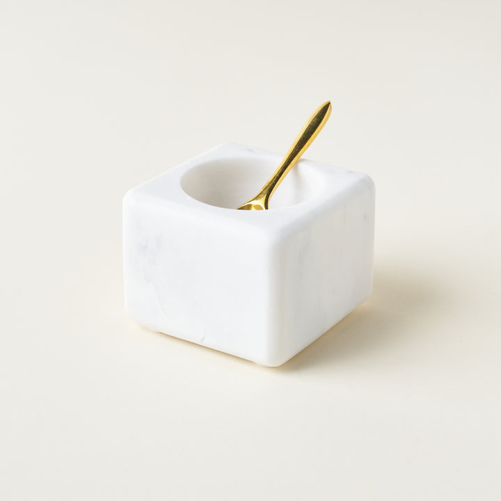 A square, white marble bowl with a rounded opening, accompanied by a polished brass spoon.