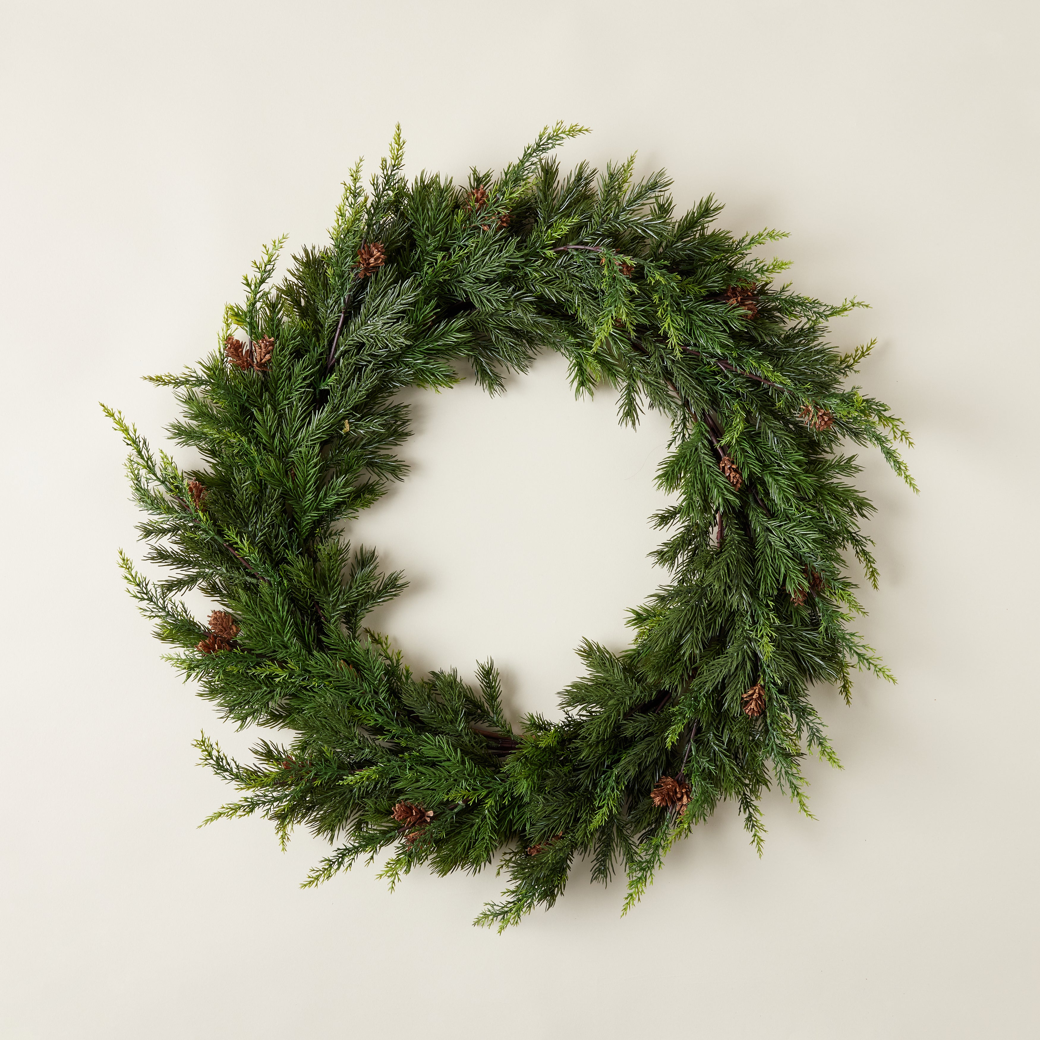 A full, round wreath made of realistic-looking faux juniper and pine branches with natural pinecones interspersed.