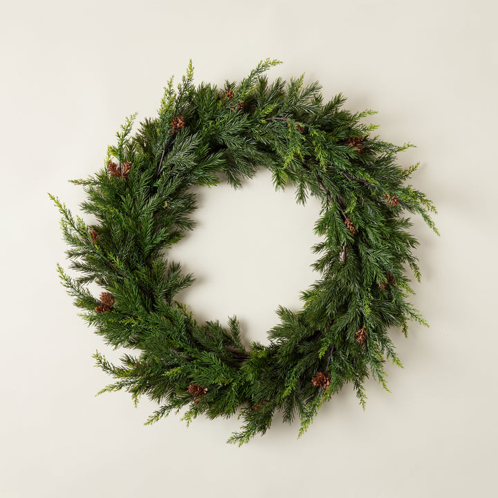 A full, round wreath made of realistic-looking faux juniper and pine branches with natural pinecones interspersed.